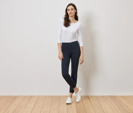 Frau steht vor einer weißen Wand, gekleidet in ein weißes Oberteil, blaue Leggings und weiße Turnschuhe.
