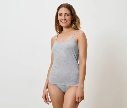 Frau mit langen braunen Haaren, die ein graues Tanktop und Unterwäsche vor einem weißen Hintergrund trägt und lächelt.