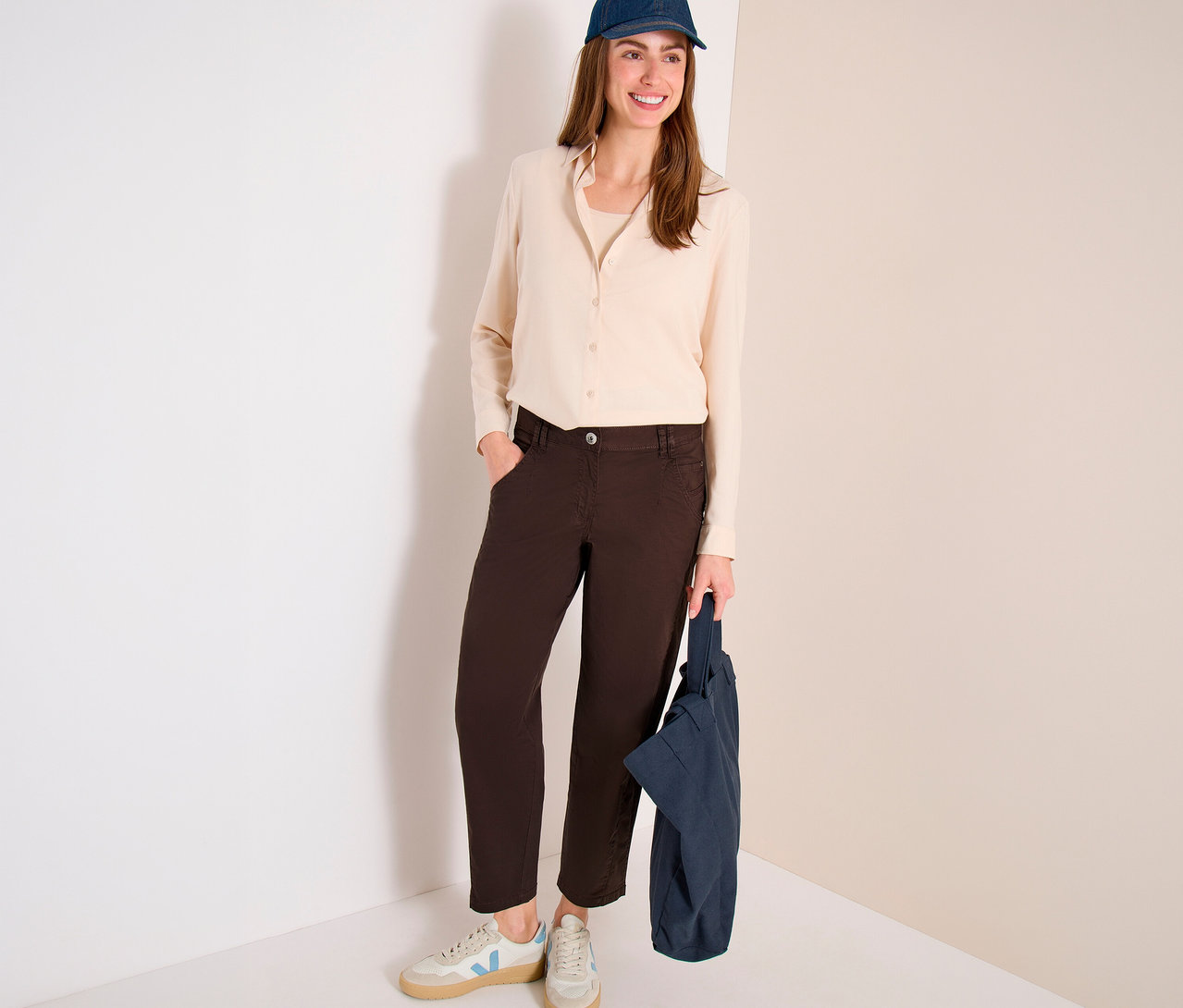 Eine lächelnde Frau steht in einem hellen Hemd, einer dunkelblauen Baseballkappe und einer braunen CECIL Barrel Hose „Scarlett“ und hält eine dunkelblaue Tragetasche.