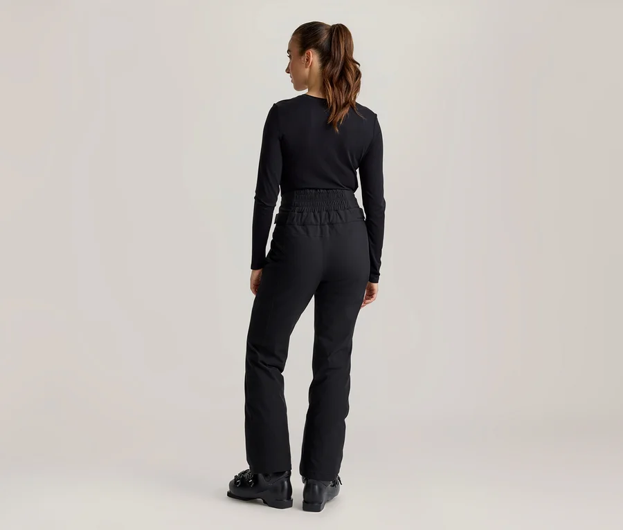 Frau von hinten, in schwarzem Langarmshirt, schwarzer Skihose mit Gummizugbund und schwarzen Skischuhen. Ihr braunes Haar ist zu einem Pferdeschwanz gebunden.