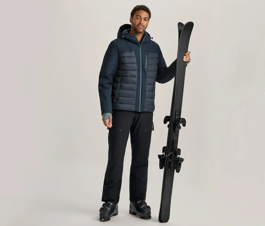 Mann in dunkelblauer Steppjacke, schwarzer Skihose und Skischuhen, der ein Paar schwarze Skier aufrecht hält.