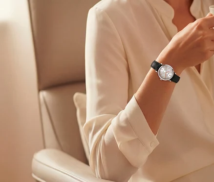 Nahaufnahme eines Arms in einer hellen Bluse, der eine silberne Uhr mit weißem Zifferblatt und schwarzem Armband trägt und ein klares Glas hält, während er in einem beigen Sessel sitzt.