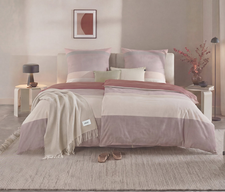 Ein Schlafzimmer mit einem Bett, das mit gestreifter SCHÖNER WOHNEN Flanell-Bettwäsche in Rosa-, Lila- und Beigetönen bezogen ist. Ein beiger Überwurf liegt auf dem Bett, daneben stehen Nachttische mit Lampen und Dekoration. Ein gestreifter Teppich liegt auf dem Boden.
