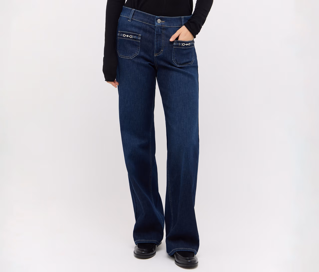 Person trägt dunkelblaue Jeans mit weitem Bein, dekorativen Vordertaschen, ein schwarzes Langarmshirt und schwarze Schuhe, eine Hand in der Tasche.