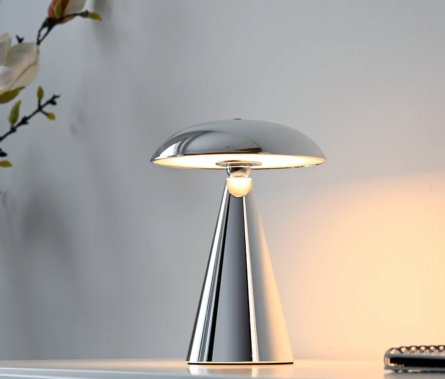 Eine verchromte Tischlampe mit pilzförmigem Schirm und konischem Fuß steht auf einer weißen Oberfläche und strahlt ein warmes Licht aus. Im Hintergrund sind eine weiße Wand und dekorative Blumen zu sehen.