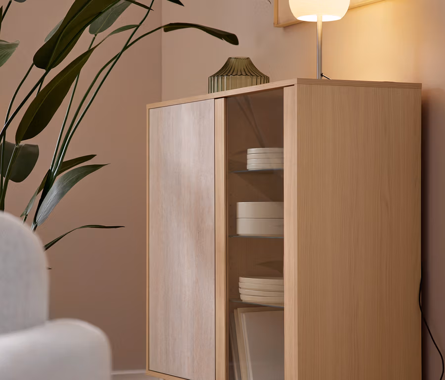 Ein heller Holzschrank mit einer Holztür und einer Glastür steht auf Parkettboden. Im Glasbereich sind weiße Teller und Schalen gestapelt. Auf dem Schrank stehen eine leuchtende weiße Kugellampe und eine grüne gerippte Vase neben einer großen Grünpflanze. Ein gerahmtes abstraktes Kunstwerk hängt an der hellbraunen Wand über der Lampe, und ein weißer Sessel ist links teilweise sichtbar.