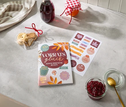 Auf einer grauen Arbeitsplatte liegen ein gestreiftes Küchentuch, eine Tüte Kekse mit roter Schleife, eine Flasche Saft und ein Glas Marmelade. Daneben ein Buch „Vorratsglück“ mit „Vielfältige Küchenetiketten und Rezepte“ und ein Bogen Aufkleber mit Sprüchen wie „Für dich“ und „Homemade“. Rechts unten steht ein offenes Glas mit roter Marmelade und einem goldenen Löffel.