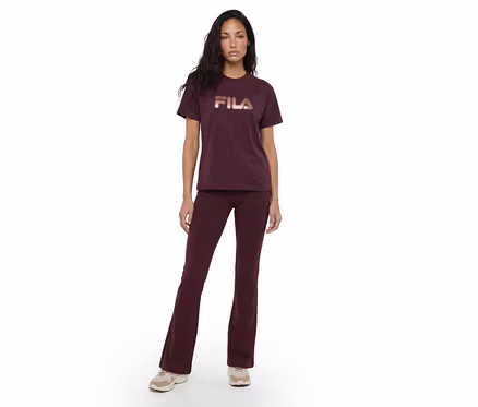Eine Frau steht frontal, trägt ein dunkelrotes T-Shirt mit roségoldenem „FILA“-Logo und dazu passende dunkelrote Schlaghosen, kombiniert mit hellen Sneakern.