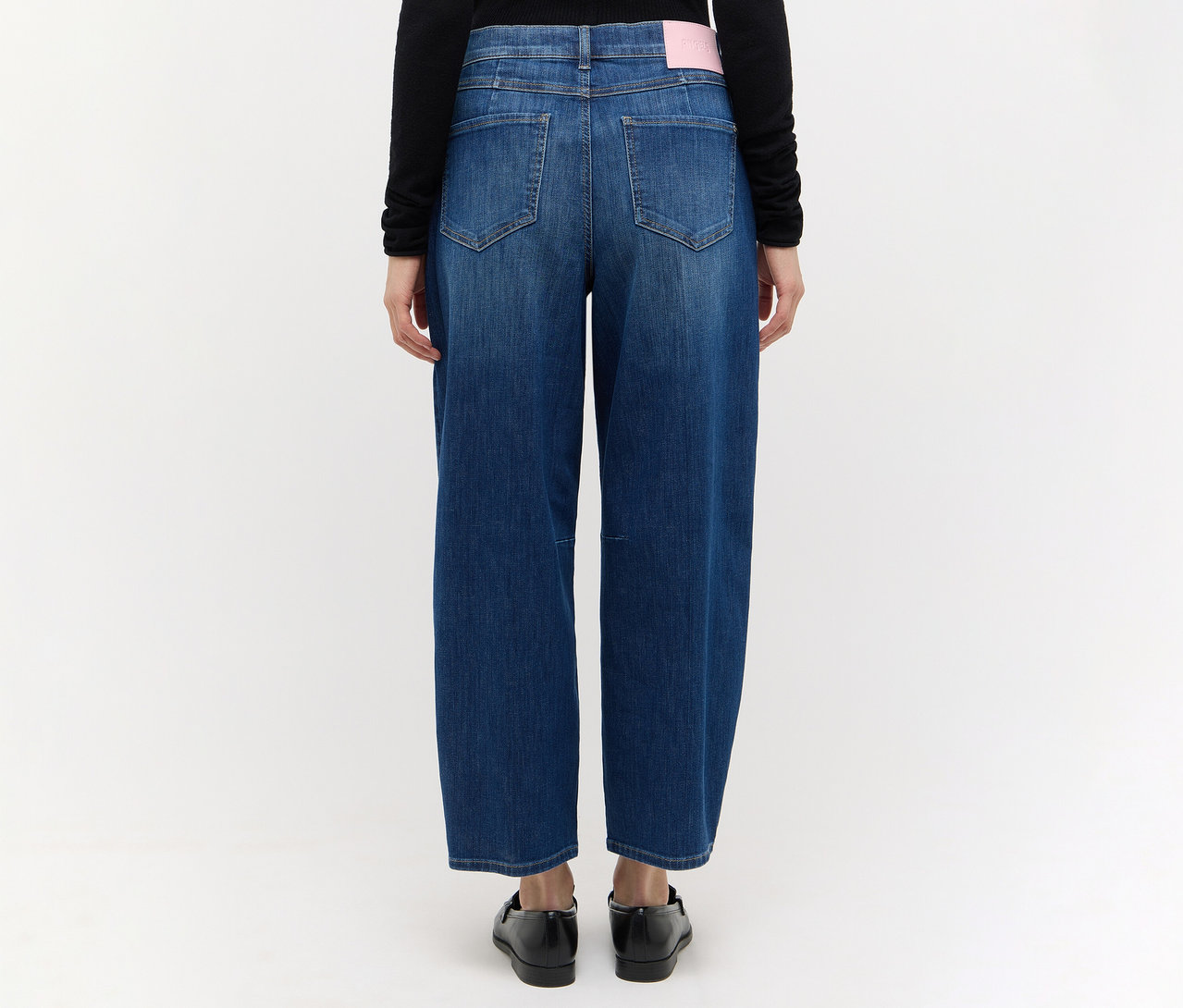 Rückenansicht einer Person in schwarzem Langarmshirt und blauer Angels Denim »Elly« Jeans mit weitem Bein und rosa Etikett. Die Person trägt schwarze Schuhe.