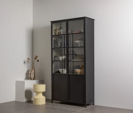 Ein hoher schwarzer Metallvitrinenschrank mit Glastüren oben und massiven Türen unten, gefüllt mit Geschirr und Dekoartikeln, steht in einer weißen Ecke. Links daneben steht ein weißer Sockel mit einer wellenförmigen Vase mit Trockenblumen, daneben ein gelber, sanduhrförmiger Hocker.
