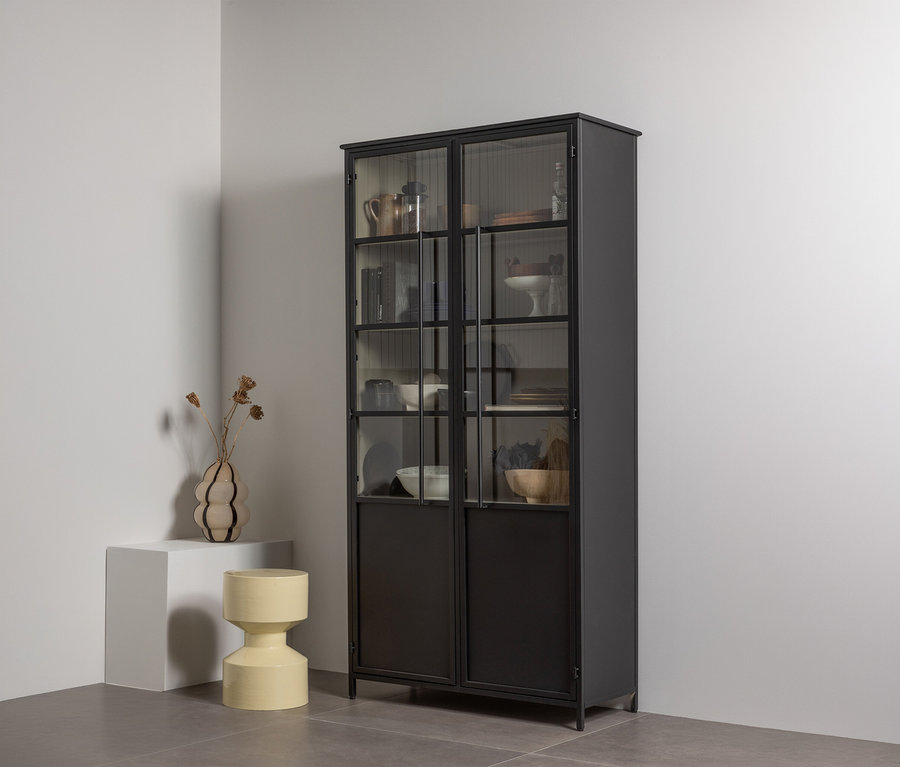 Ein hoher schwarzer Metallvitrinenschrank mit Glastüren oben und massiven Türen unten, gefüllt mit Geschirr und Dekoartikeln, steht in einer weißen Ecke. Links daneben steht ein weißer Sockel mit einer wellenförmigen Vase mit Trockenblumen, daneben ein gelber, sanduhrförmiger Hocker.