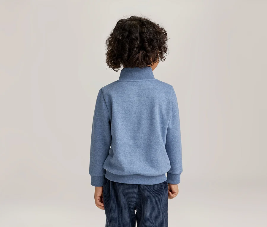 Kind von hinten gesehen, trägt ein blaues Sweatshirt mit Stehkragen und eine dunkelblaue Cordhose. Das Kind hat lockiges, dunkles Haar.