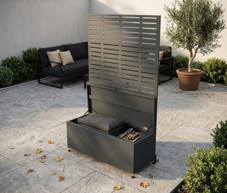 Dunkelgraue Outdoor-Aufbewahrungsbox mit offenem Deckel, der Kissen und Gartengeräte zeigt, und einem integrierten Lamellen-Sichtschutz, auf einer Steinterrasse mit Outdoor-Sofa und Olivenbaum im Hintergrund.