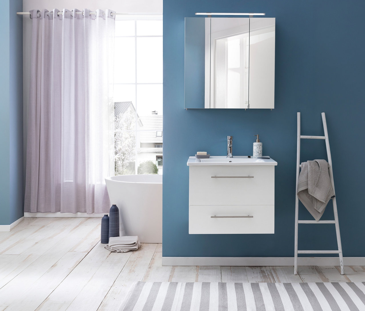 Modernes Badezimmer mit blauen Wänden und hellem Holzboden. Über einem weißen Waschtisch hängt ein Spiegelschrank »Dorina« mit Licht, 60 cm, mit drei Spiegeltüren. Links steht eine freistehende Badewanne vor einem Fenster mit Vorhängen, rechts ein weißer Handtuchhalter mit einem grauen Handtuch. Auf dem Holzboden liegen ein gestreifter Teppich, zwei blaue Vasen und ein gefaltetes Handtuch.