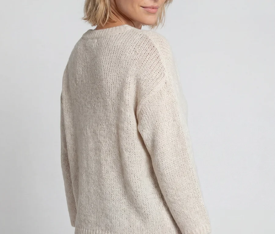 Frau mit kurzem blondem Haar, die einen beigefarbenen Olsen 3/4-Pullover und olivgrüne Hosen trägt und über ihre rechte Schulter in die Kamera blickt.
