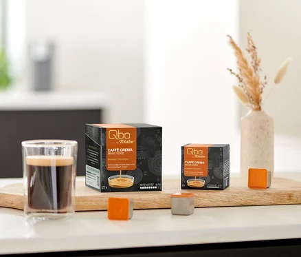 Zwei Qbo Caffè MINAS VERDE Kapselpackungen, ein Glas Kaffee und lose Kapseln auf einem Holzbrett auf der Küchenzeile, daneben Kekse und Trockenblumen.