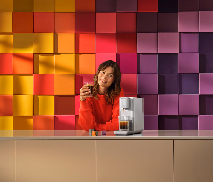Lächelnde Frau in rotem Oberteil hält ein Glas Kaffee, während eine silberne Kaffeemaschine Kaffee in ein Glas füllt. Bunter Hintergrund aus quadratischen Blöcken.