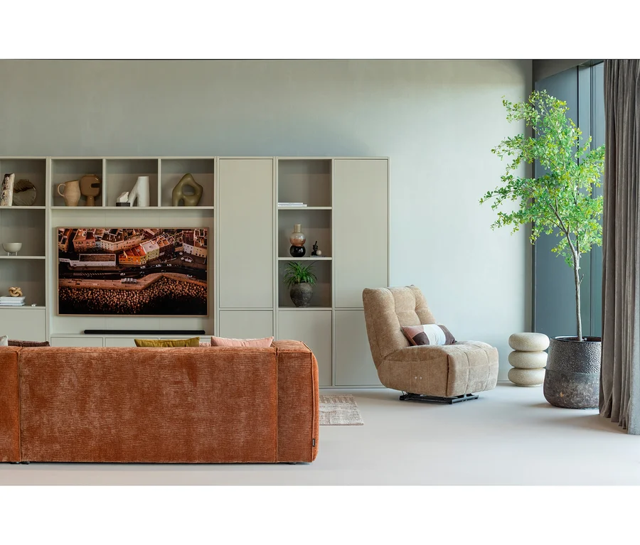 Ein modernes Wohnzimmer mit einem orangefarbenen strukturierten Sofa, einem beigefarbenen Polstersessel und einer hellgrünen Wohnwand. Die Wohnwand zeigt einen Fernseher mit einer Stadtszene und diverse Deko-Objekte. Eine hohe grüne Pflanze in einem dunklen Topf steht neben einem Fenster mit dunklen Vorhängen, daneben drei gestapelte helle Hocker.