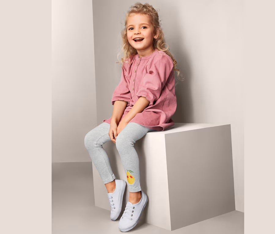 Ein lächelndes blondes Mädchen sitzt auf einem weißen Würfel und trägt ein rosa Kleid und 2 Leggings mit Zitronenprint und weiße Turnschuhe.