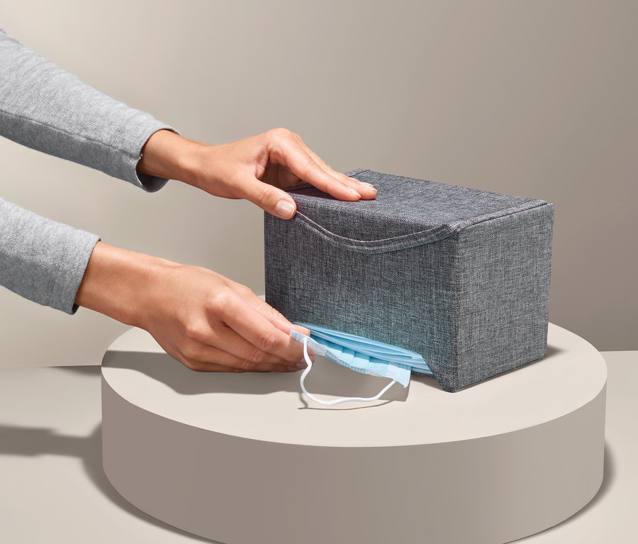 Hände entnehmen eine blaue Maske aus einer grauen Maskenbox.