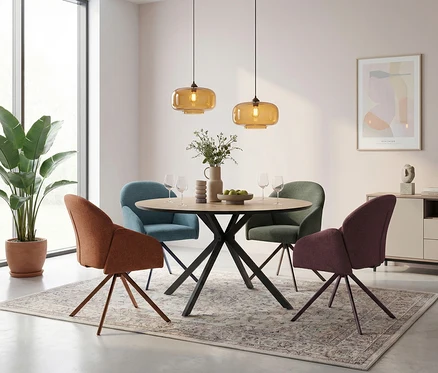 Runder Esstisch mit vier Stühlen in verschiedenen Farben (Orange, Blau, Grün, Violett) auf einem gemusterten Teppich. Über dem Tisch hängen zwei bernsteinfarbene Pendelleuchten. Auf dem Tisch stehen eine Vase mit Grünzeug, Weingläser und eine Obstschale. Im Hintergrund sind ein großes Fenster mit einer Pflanze, ein Sideboard und ein gerahmtes Bild zu sehen.