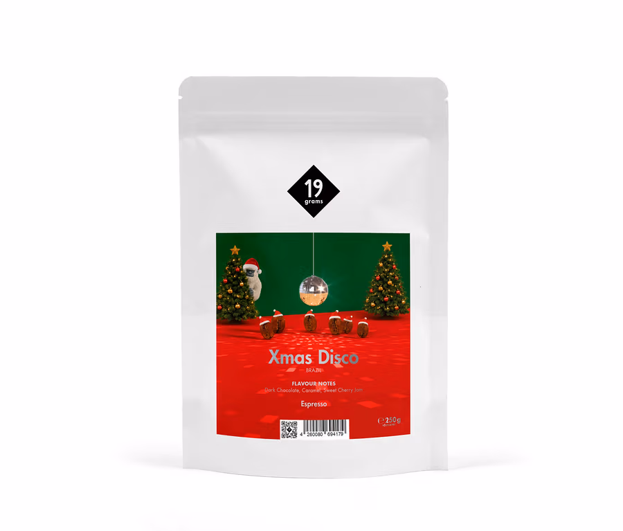 Weißer Kaffeebeutel mit rotem Etikett, das eine Weihnachts-Disco-Szene zeigt: zwei geschmückte Christbäume, ein Faultier mit Weihnachtsmütze, eine Discokugel und kleine braune Kuchen auf rotem Boden vor einer grünen Wand. Das Etikett trägt das „19 grams“-Logo, „Xmas Disco Brazil“, „Flavour Notes: Dark Chocolate, Caramel, Sweet Cherry Jam“, „Espresso“ und „250g“.