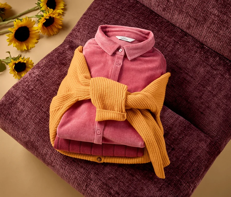 Ein gefaltetes pinkes Cordhemd und ein dunkelpinkes Rippentop, um die ein gelber Merino-Cardigan gebunden ist, liegen auf einer dunkelroten, strukturierten Oberfläche. Im Hintergrund sind Sonnenblumen zu sehen.