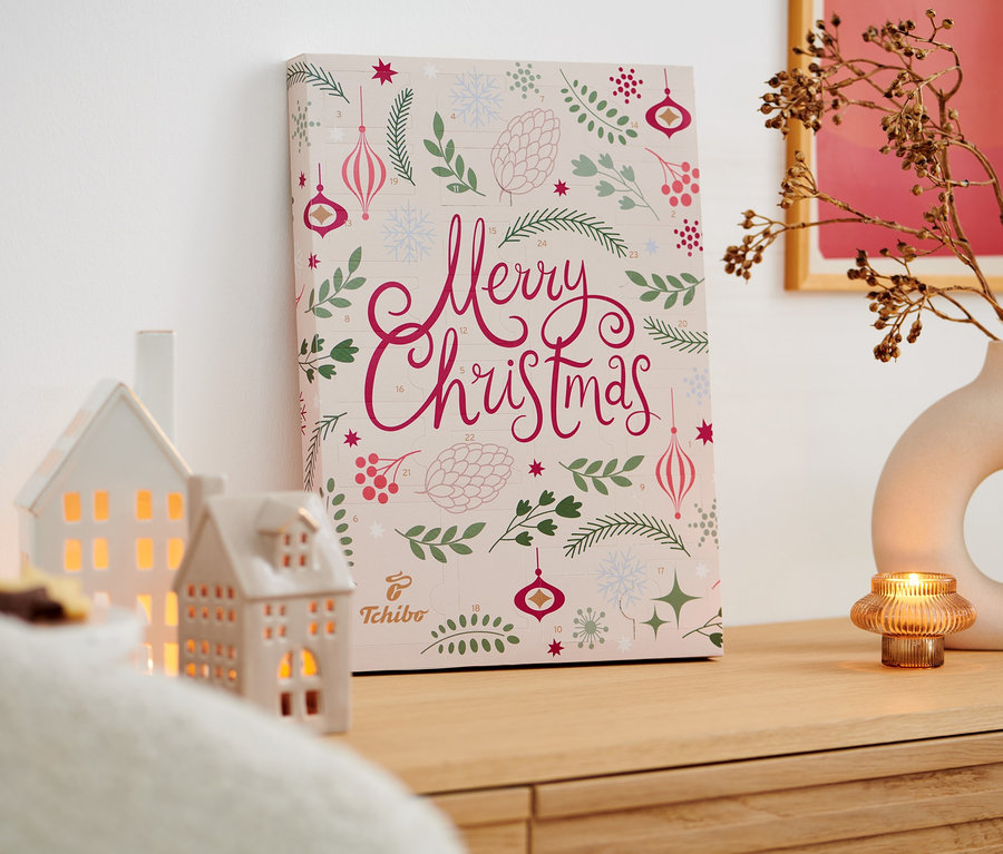 Ein Tchibo-Adventskalender steht auf einer Holzkommode neben zwei Häusern, einer Vase mit Zweigen und einer brennenden Kerze.