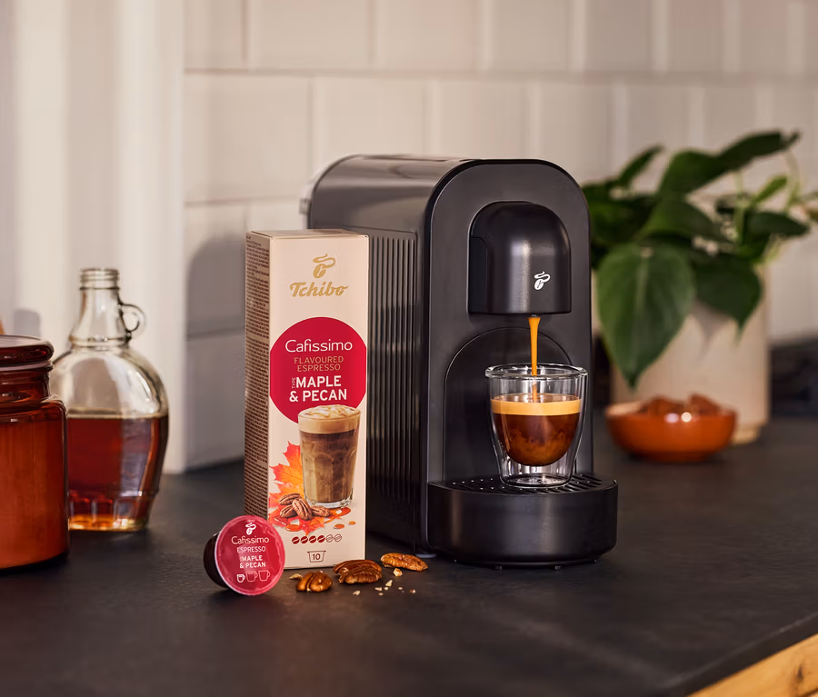Schwarze Cafissimo now Kaffeemaschine brüht Cafissimo Flavoured Espresso Maple & Pecan in ein Glas. Daneben Kapselpackung, lose Kapsel, Nüsse, Flasche und Pflanze.