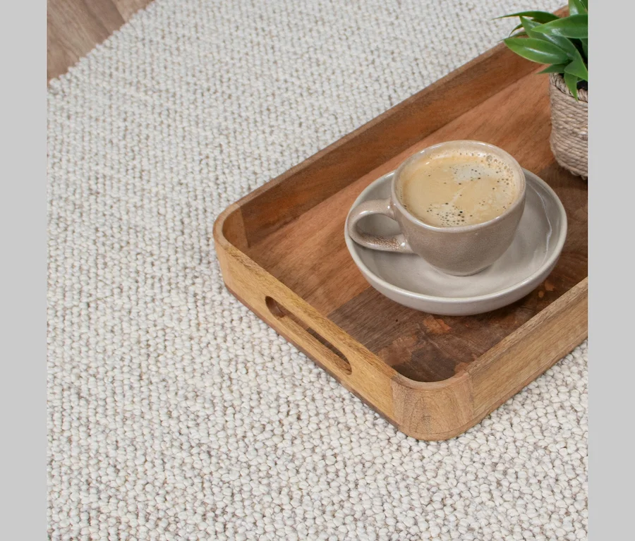 Holztablett mit einer Tasse Kaffee und Untertasse auf einem hellen, strukturierten Teppich, daneben eine kleine Pflanze in einem geflochtenen Topf.