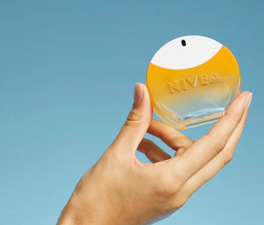 Eine Hand hält eine runde Nivea Flasche mit klarem Boden, gelber Flüssigkeit und weißer Kappe vor blauem Hintergrund.