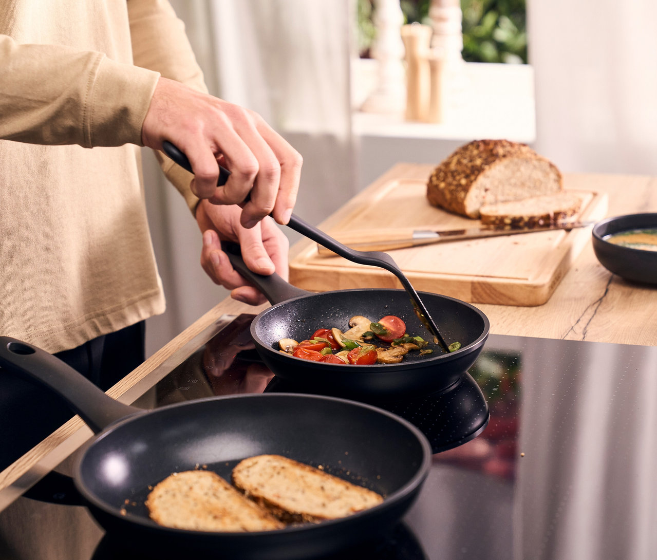 Eine Person rührt Gemüse in einer Henckels by ZWILLING Breeze Bratpfanne; daneben toastet in einer weiteren Henckels by ZWILLING Breeze Bratpfanne Brot. Im Hintergrund liegen ein Brotlaib und ein Messer auf einem Holzbrett, eine Schale Suppe ist ebenfalls sichtbar.
