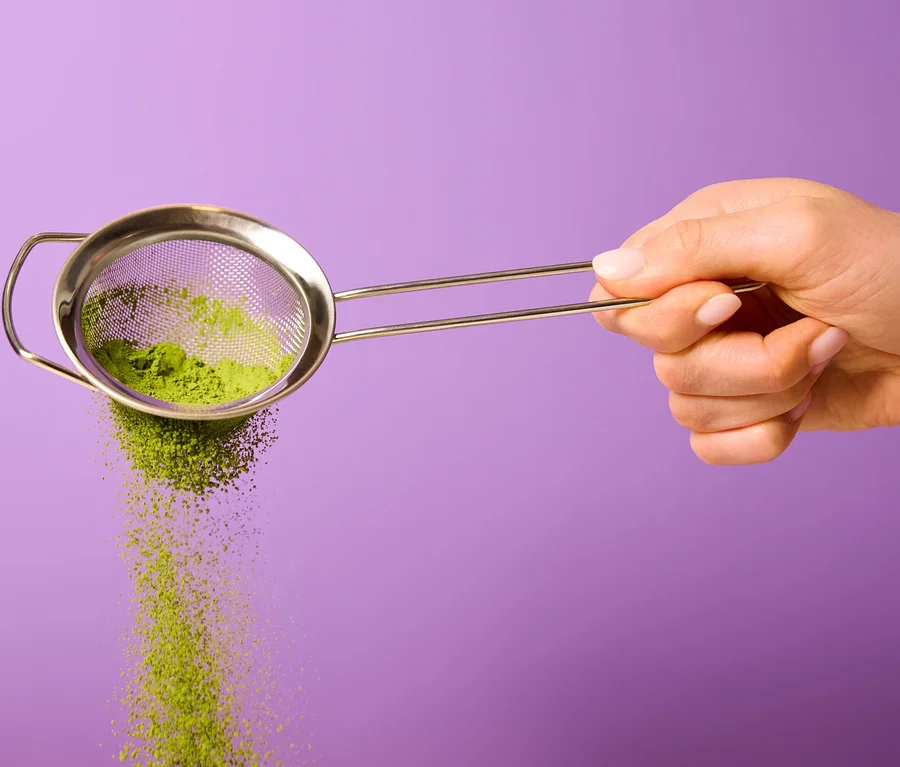 Eine Hand hält ein Metallsieb, aus dem grünes Bio Matcha by Tchibo Pulver auf einem lila Hintergrund fällt.