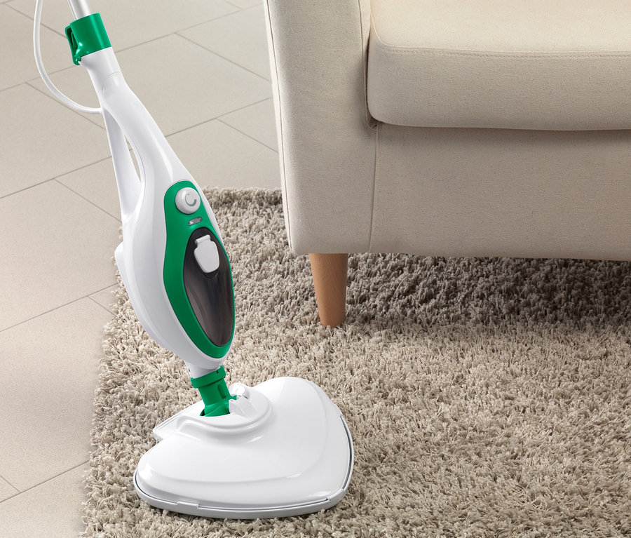 Multifunktionaler Dampfbesen auf beigem Teppich neben cremefarbenem Sessel.