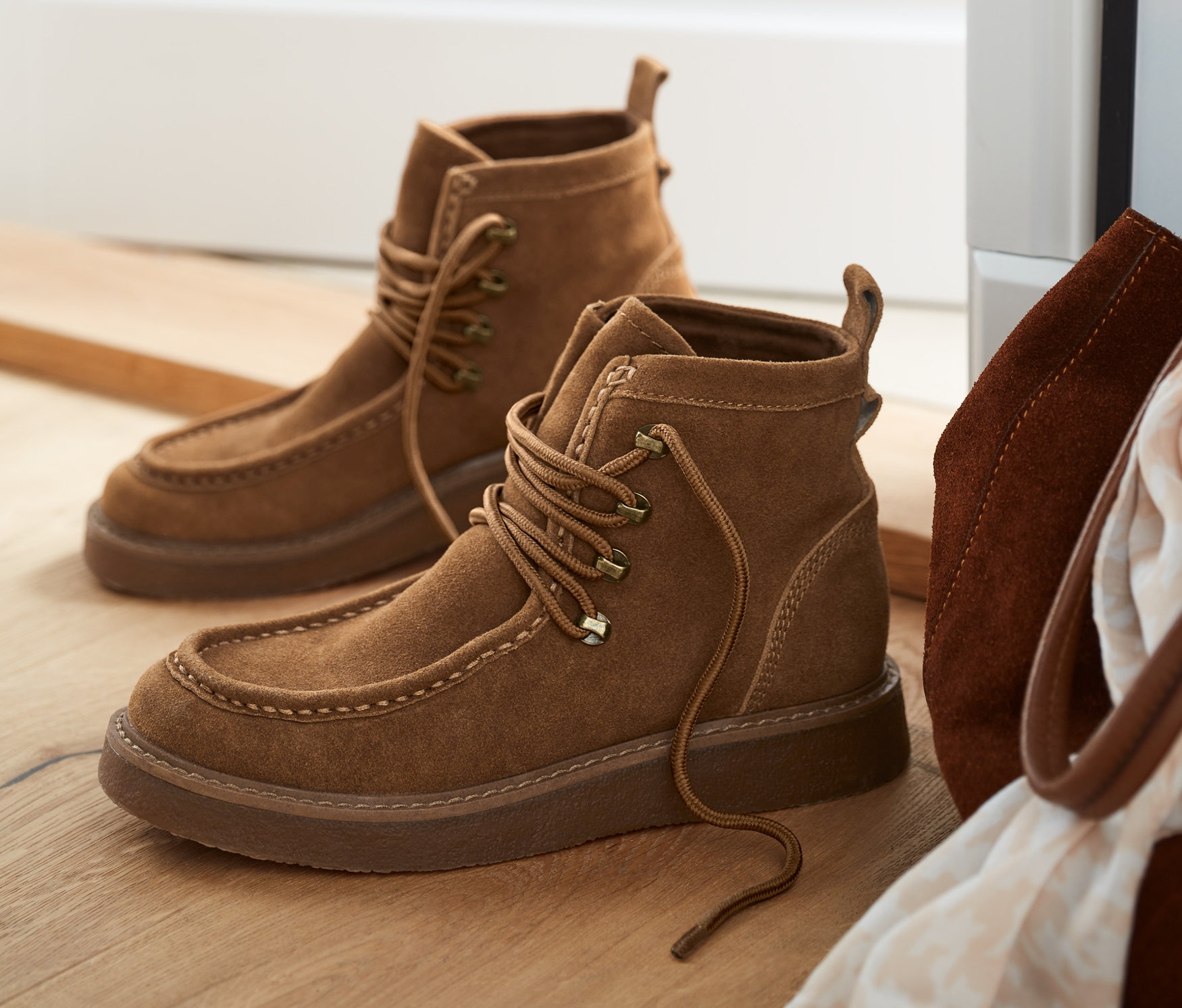 Zwei Paar Veloursleder-Boots liegen auf einem Holzboden neben einer braunen Ledertasche und einem weißen Schal.