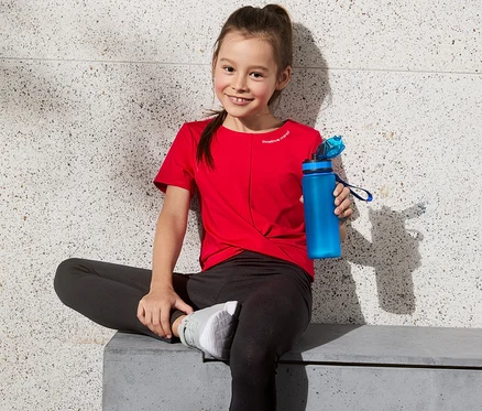 Mädchen sitzt auf Bank in rotem Cropped-Funktionsshirt mit recyceltem Material und hält blaue Wasserflasche. Basketball daneben.