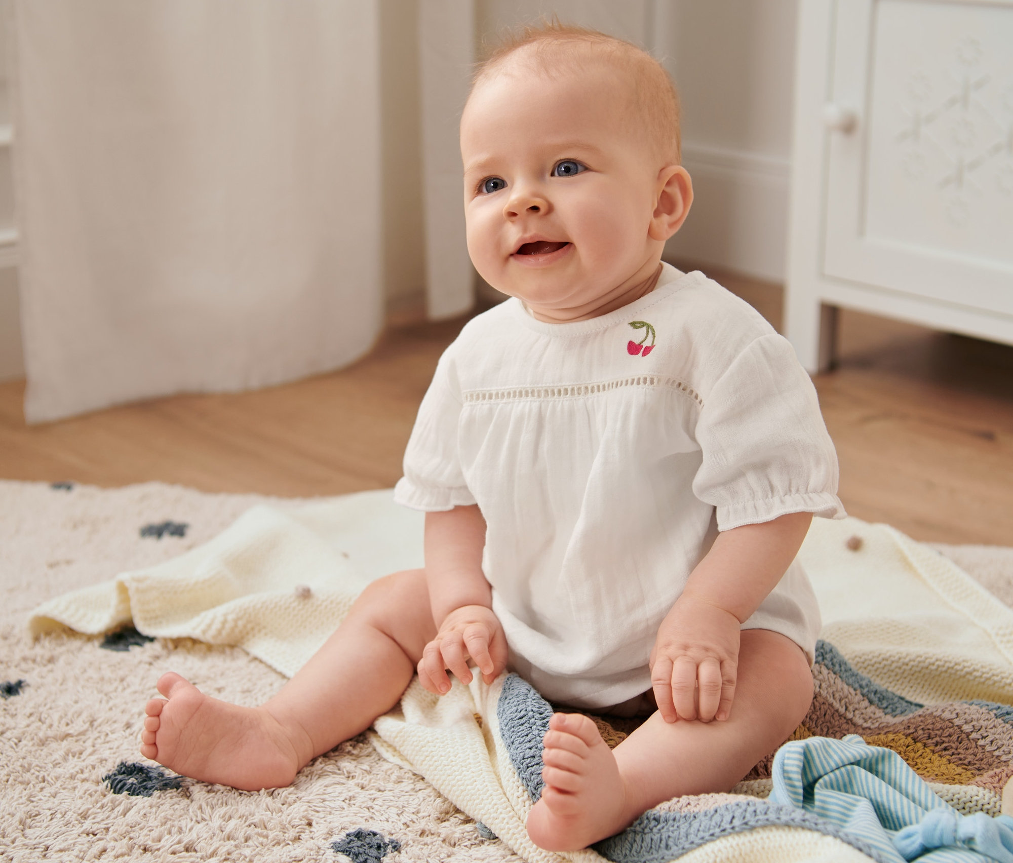 Lächelndes Baby sitzt auf einer Decke und trägt eine Bluse.