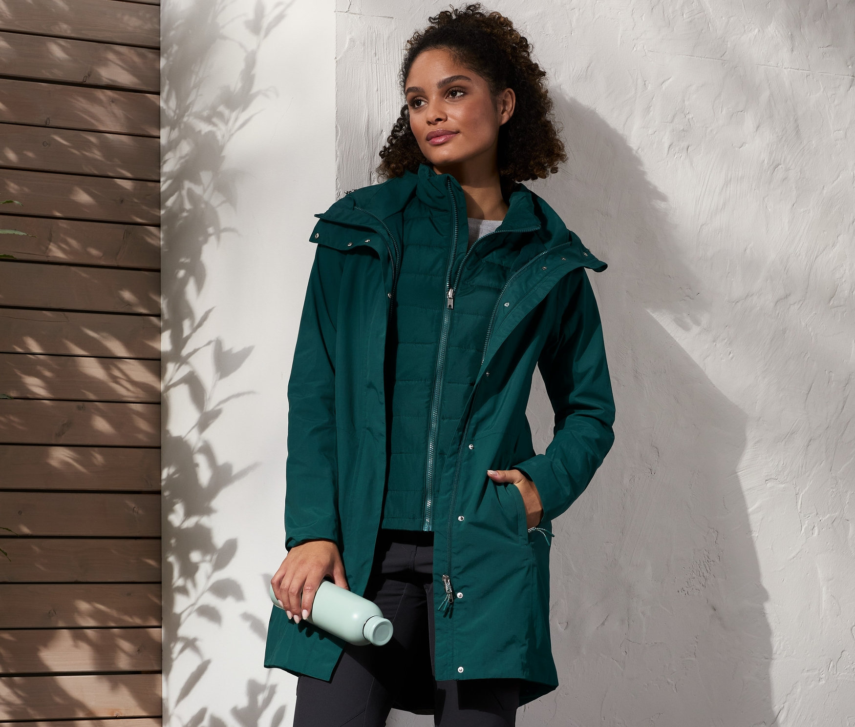 Eine Frau steht und trägt einen grünen Regenmantel mit Weste, schwarze Hosen und braune Stiefel. Sie hält eine Wasserflasche in der Hand, neben ihr steht ein Rucksack.