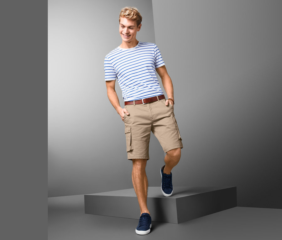 Mann posiert auf grauem Podest in Cargo-Bermudas, gestreiftem T-Shirt und Turnschuhen.