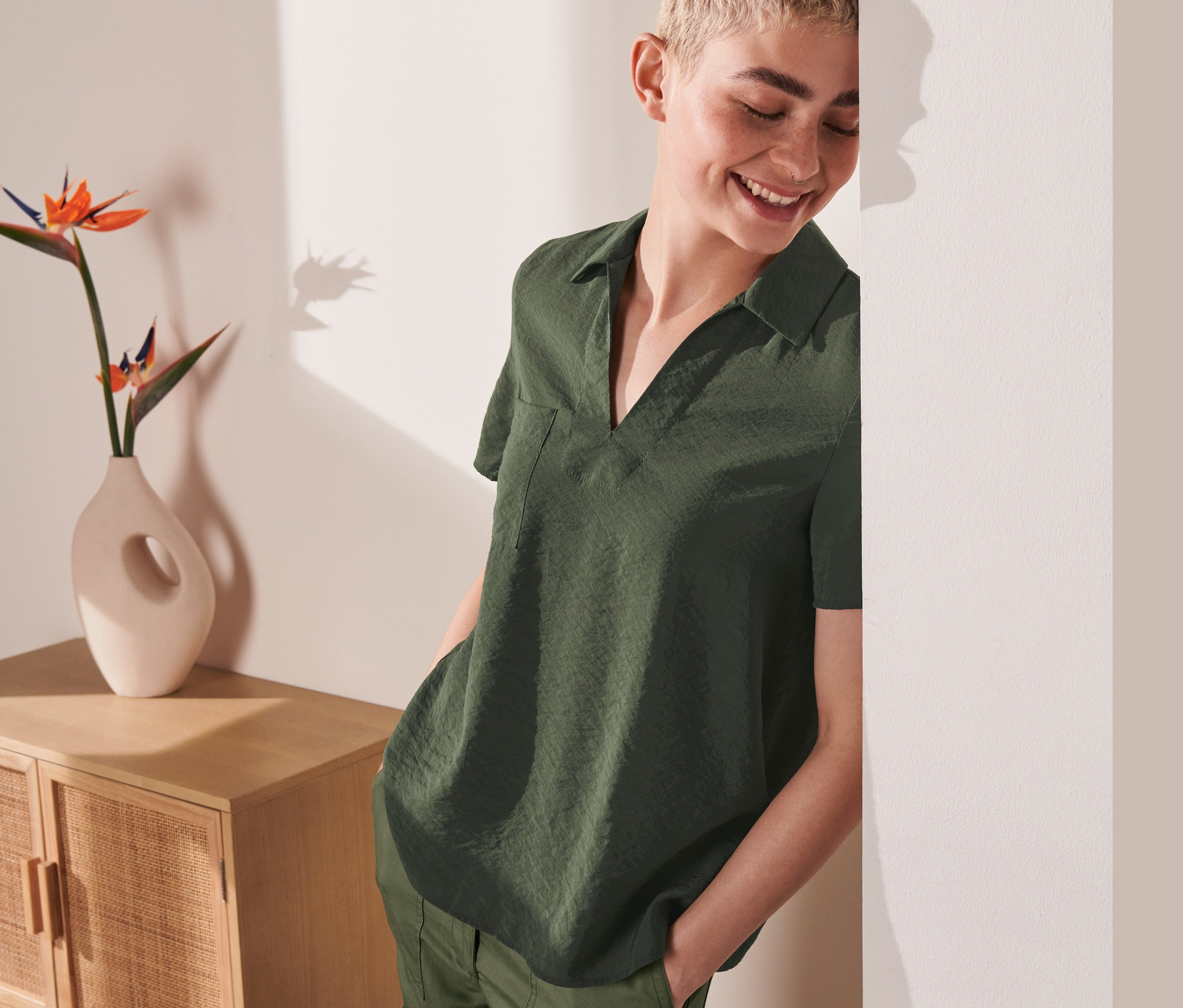 Lächelnde, kurzhaarige Frau lehnt an einer Wand und trägt eine grüne Bluse.