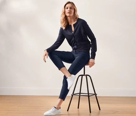 Blonde Frau sitzt auf einem Hocker und trägt eine dunkelblaue Stretchhose und eine Strickjacke.