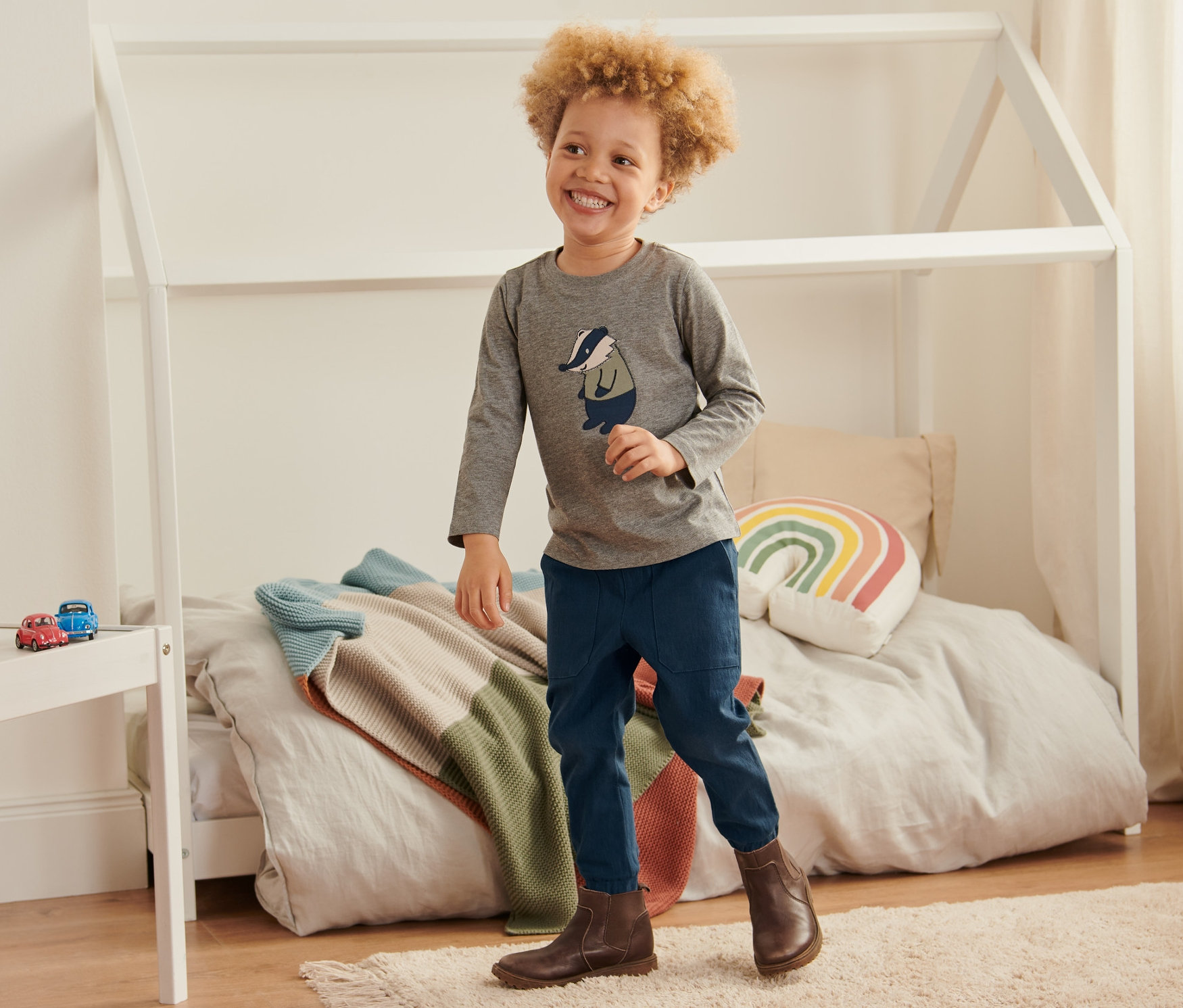 Ein lächelnder Junge steht in einem Kinderzimmer auf einem Teppich. Er trägt ein graues Langarmshirt und eine blaue Schlupfhose.