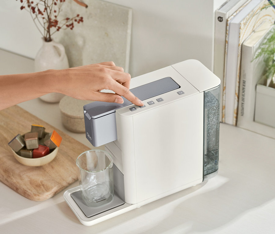 Frau bedient Qbo ICONIC Dark Chrome und Qbo ESSENTIAL Bright Sand Kaffeemaschine. Eine Schale mit Kaffeekapseln steht daneben.