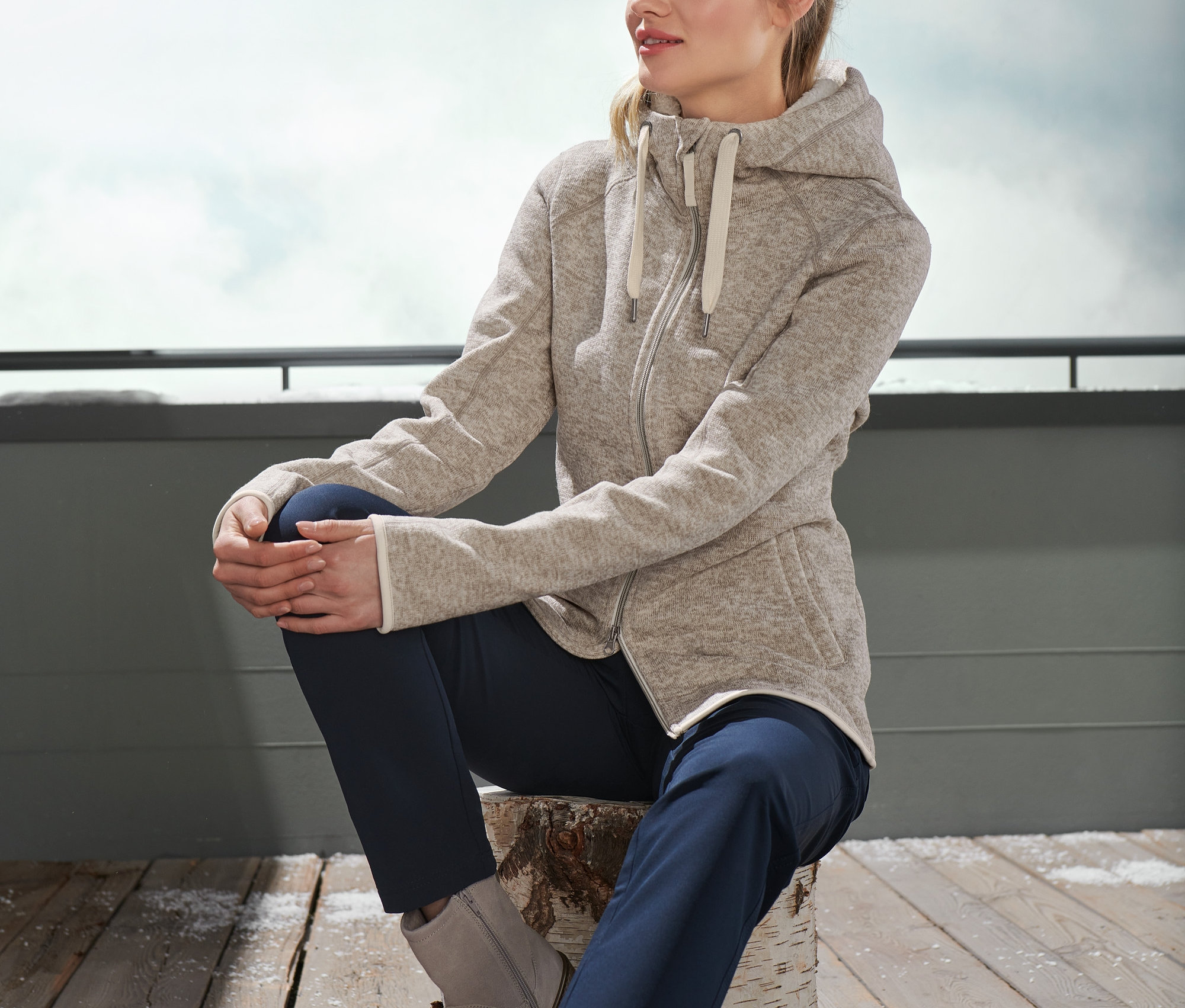 Frau sitzt auf einem Holzscheit und trägt eine beige Teddy-Strickfleecejacke.