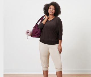 Frau mit Afro-Frisur trägt eine Tasche mit Yogamatte, trägt ein Schokoladen-Yogashirt und eine beige 3/4-Yogahose.