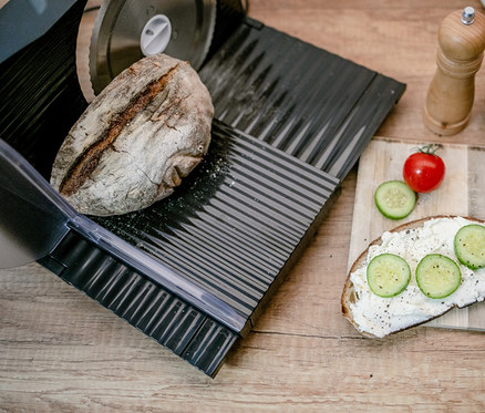 Brot liegt auf dem Steba Elektrischer Allesschneider »AS 100«. Daneben liegen Brot mit Aufstrich, Gurke und Tomate.