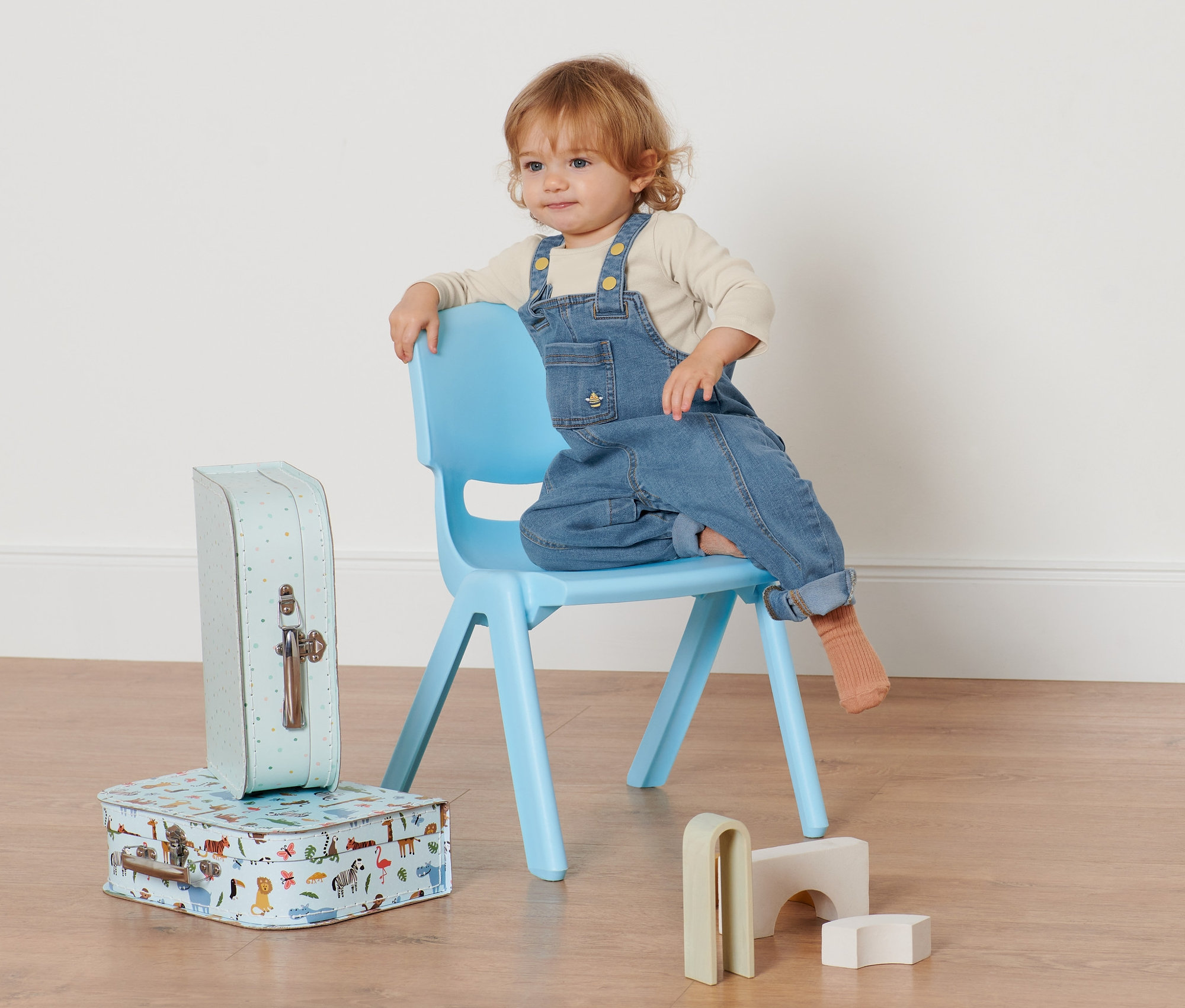 Kleinkind sitzt auf einem blauen Stuhl und trägt eine Denim-Latzhose. Neben dem Stuhl stehen zwei Koffer und Holzspielzeug.