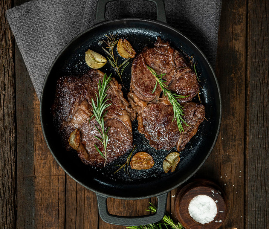 Zwei Steaks mit Rosmarin und Knoblauch in der COMBEKK Gusseisen-Servierpfanne »Sous-Chef« auf einem Holztisch.