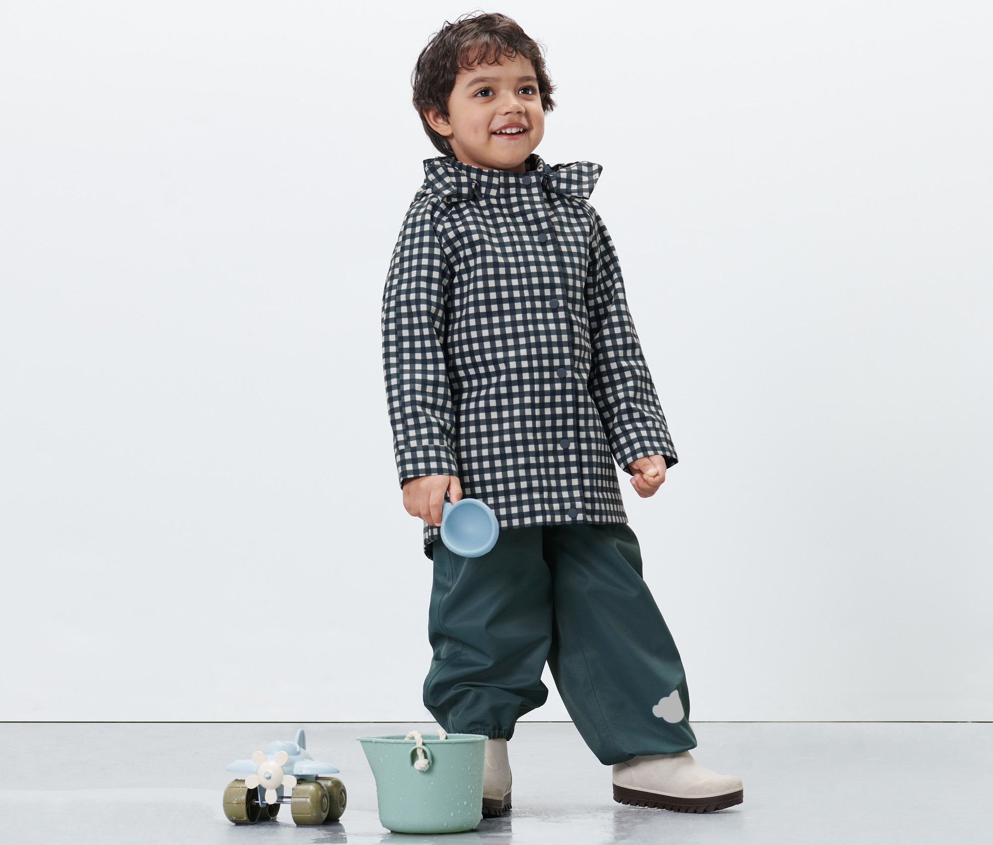 Ein Junge in einer karierten Kleinkind-Regenjacke hält eine blaue Tasse. Unten stehen ein grüner Eimer und ein Spielzeug.