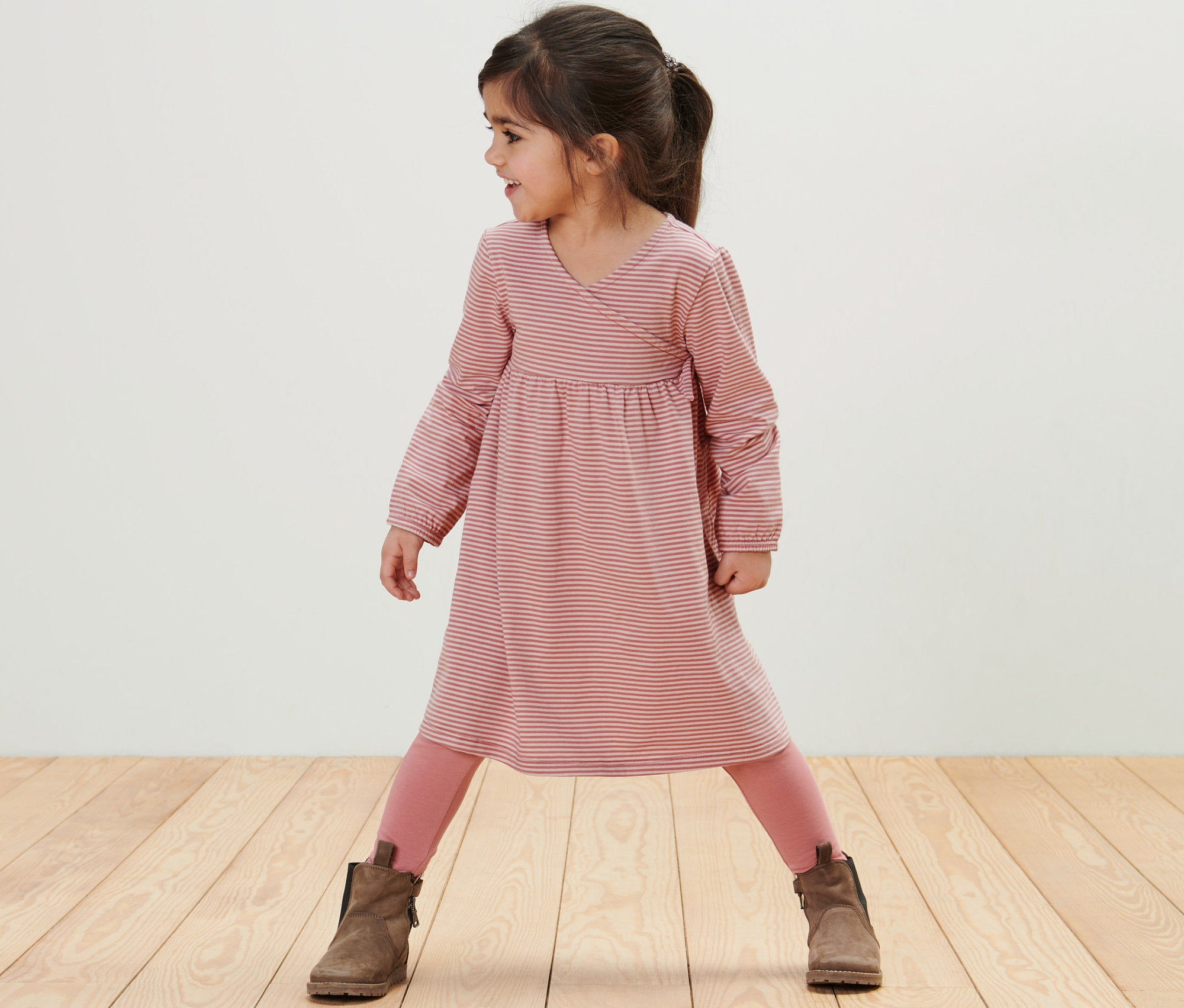Mädchen steht auf einem Holzboden und trägt ein Kinder-Jerseykleid mit rosa Leggings und braunen Stiefeln.